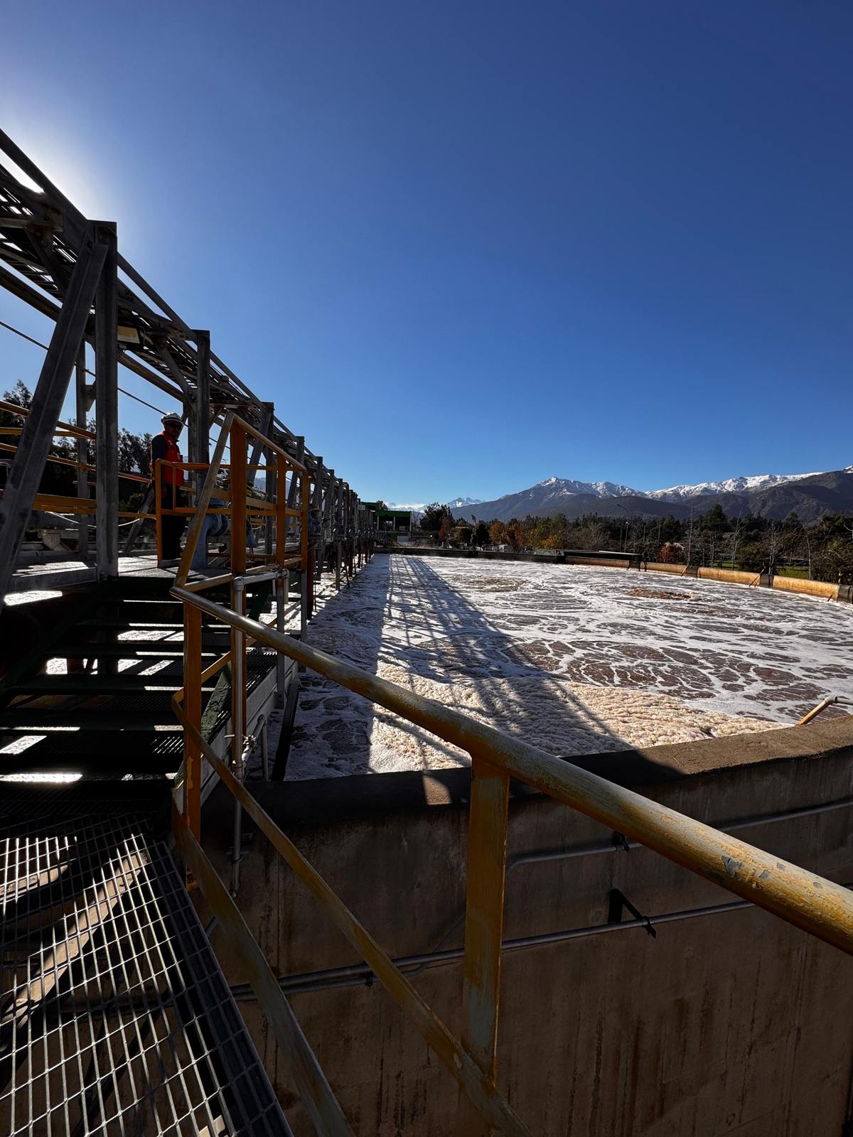Estanque de una planta de tratamiento con una pasarela metálica y la cordillera nevada al fondo